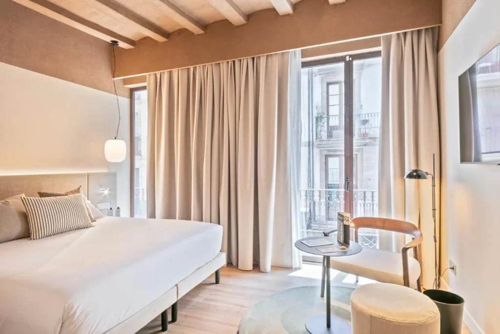 Habitaci&oacute;n moderna con cama, l&aacute;mpara y cortinas beige frente a ventanales