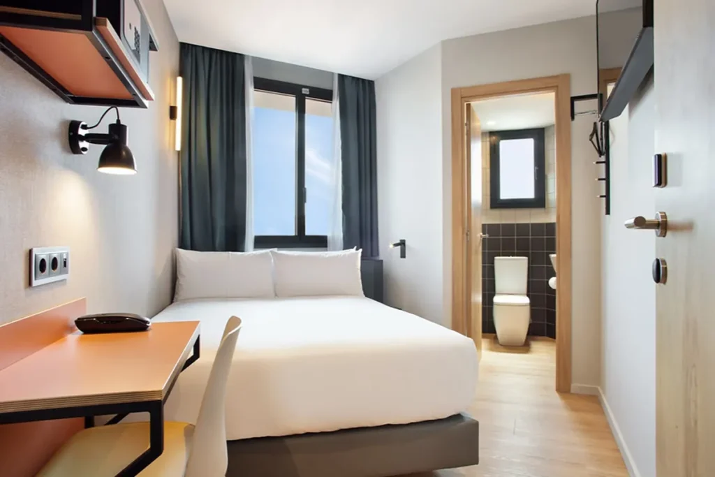 Habitaci&oacute;n moderna con cama, cortinas oscuras y ba&ntilde;o con inodoro al fondo.