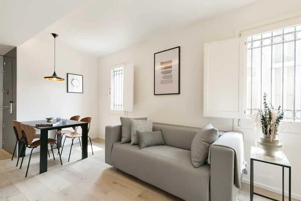 Sala luminosa con sof&aacute; gris, l&aacute;mpara, mesa y flores junto a la ventana