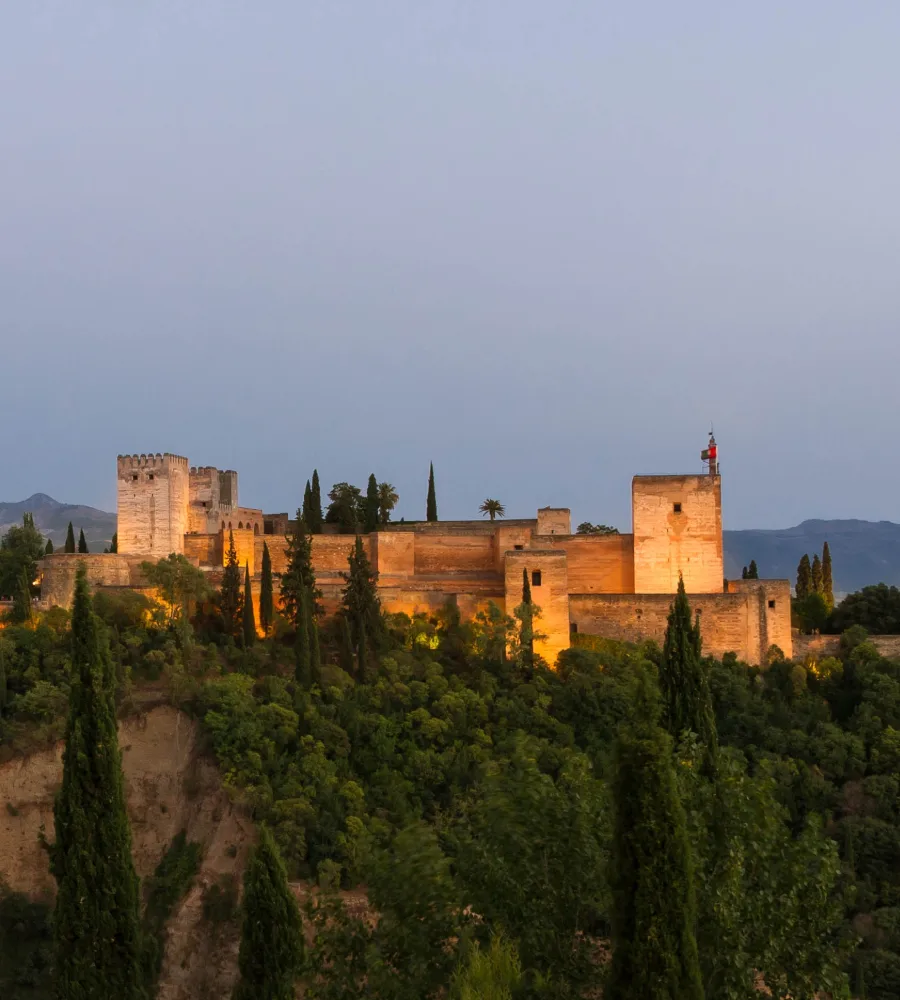 Fortaleza de piedra con torre y faro, al atardecer, entre &aacute;rboles y monta&ntilde;as