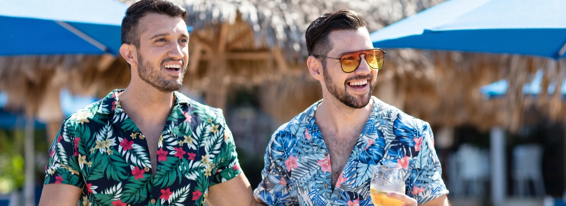 Two men in floral shirts smiling outdoors under blue umbrellas