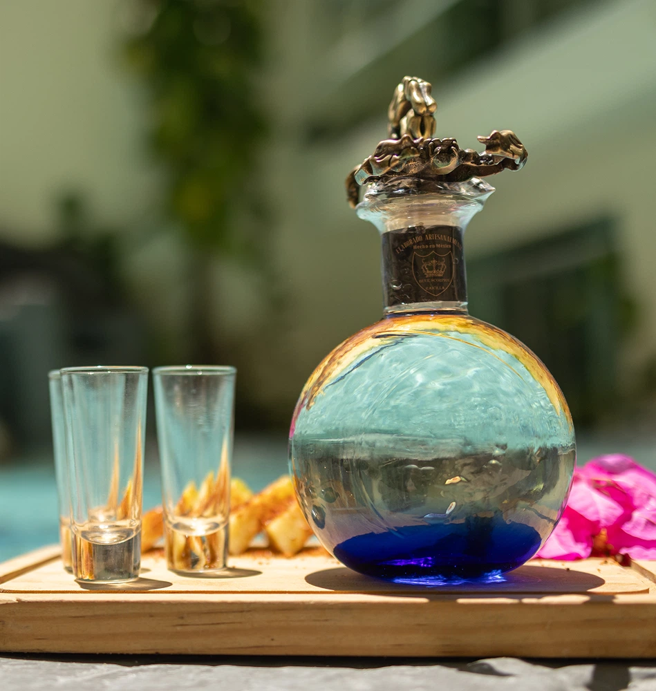 Colorful glass bottle with gold cap, two clear glasses, and blurred outdoor background