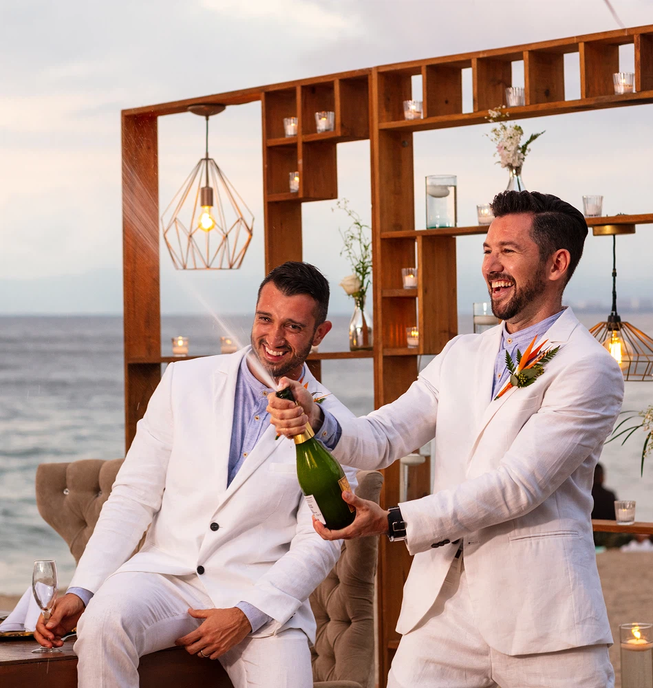 Two men in white suits toasting champagne on a beach wedding backdrop