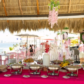 Outdoor banquet table with pink floral decor and buffet under straw roof