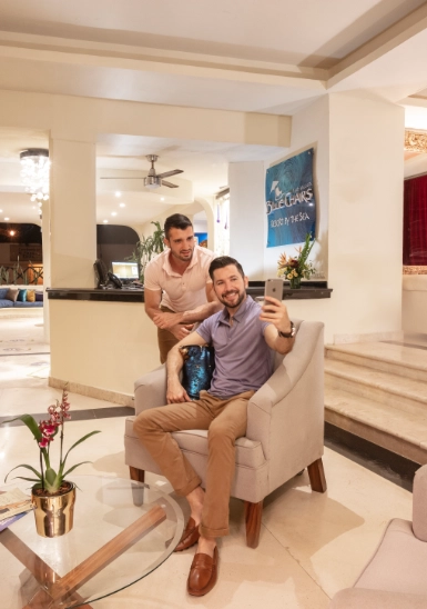 Two men smiling and posing on a sofa indoors, holding drinks.