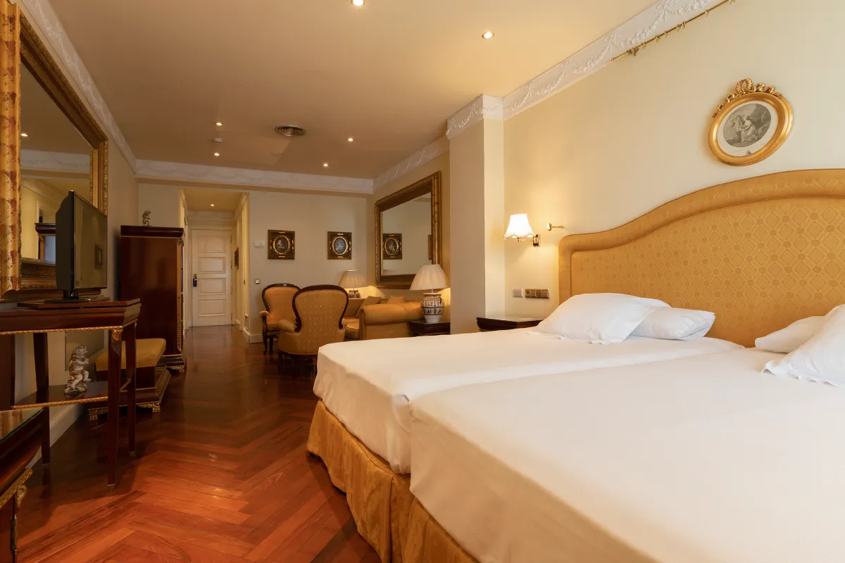 Suite hotelera con cama doble y lámparas, sala al fondo y decoración clásica.