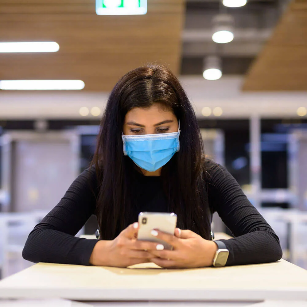 Mujer en oficina usando mascarilla y tel&eacute;fono m&oacute;vil.