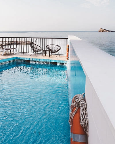 Piscina azul junto al mar, con tumbona y barandilla blanca.