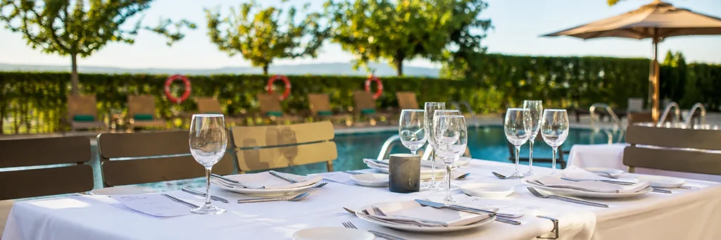 Mesa puesta al aire libre junto a piscina, copas y cubiertos sobre manteles blancos