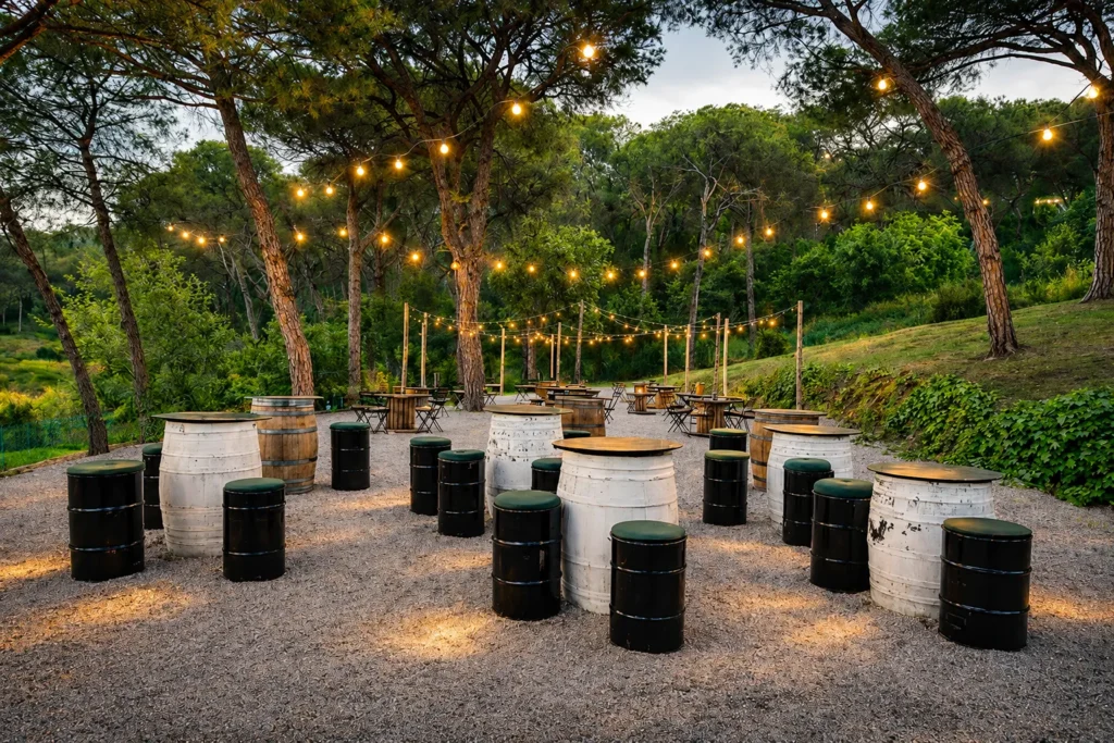 Espacio al aire libre con barriles y luces colgantes entre &aacute;rboles.