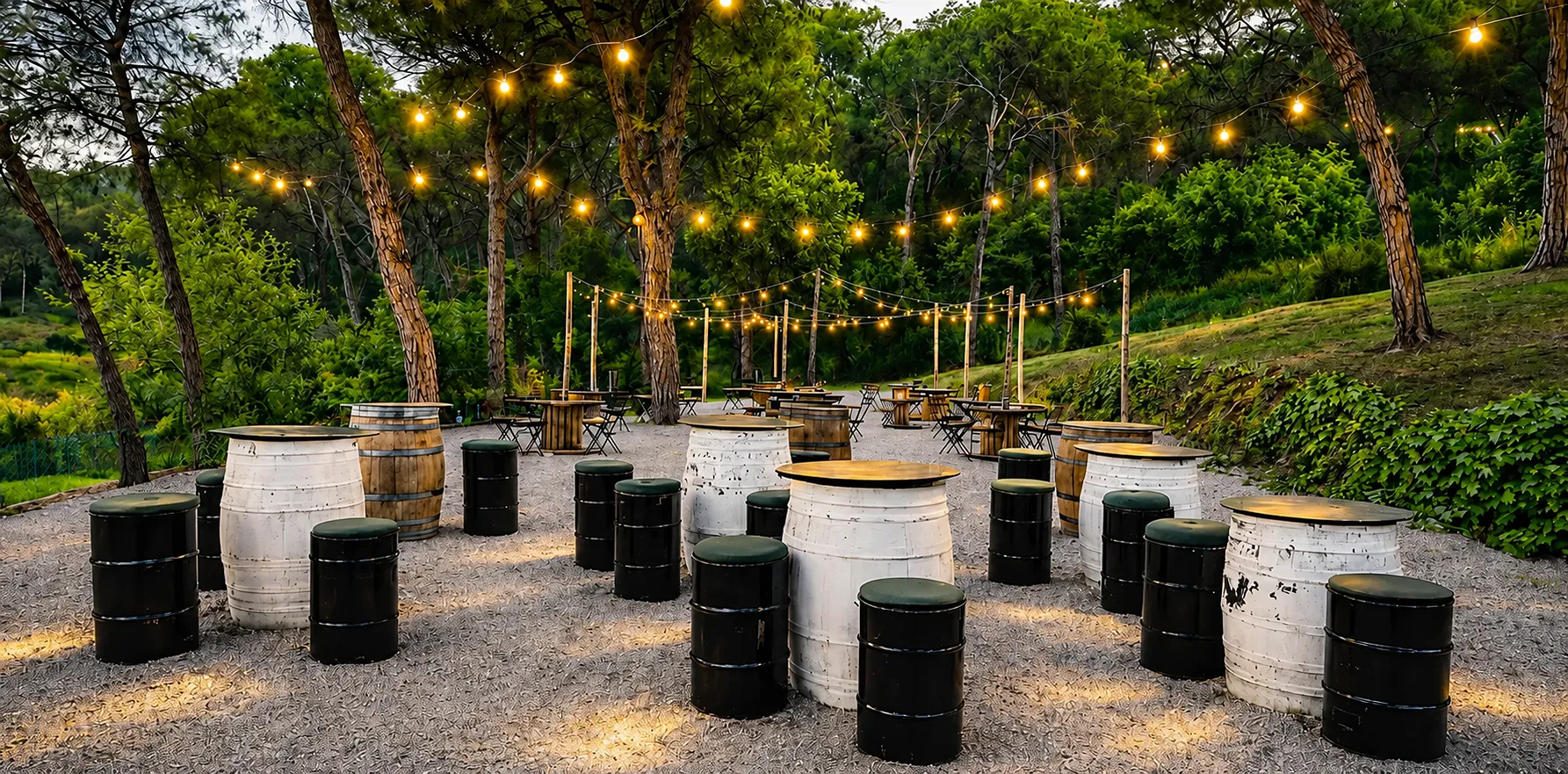 Patio verde con barriles y mesas, guirnalda de luces colgante al atardecer