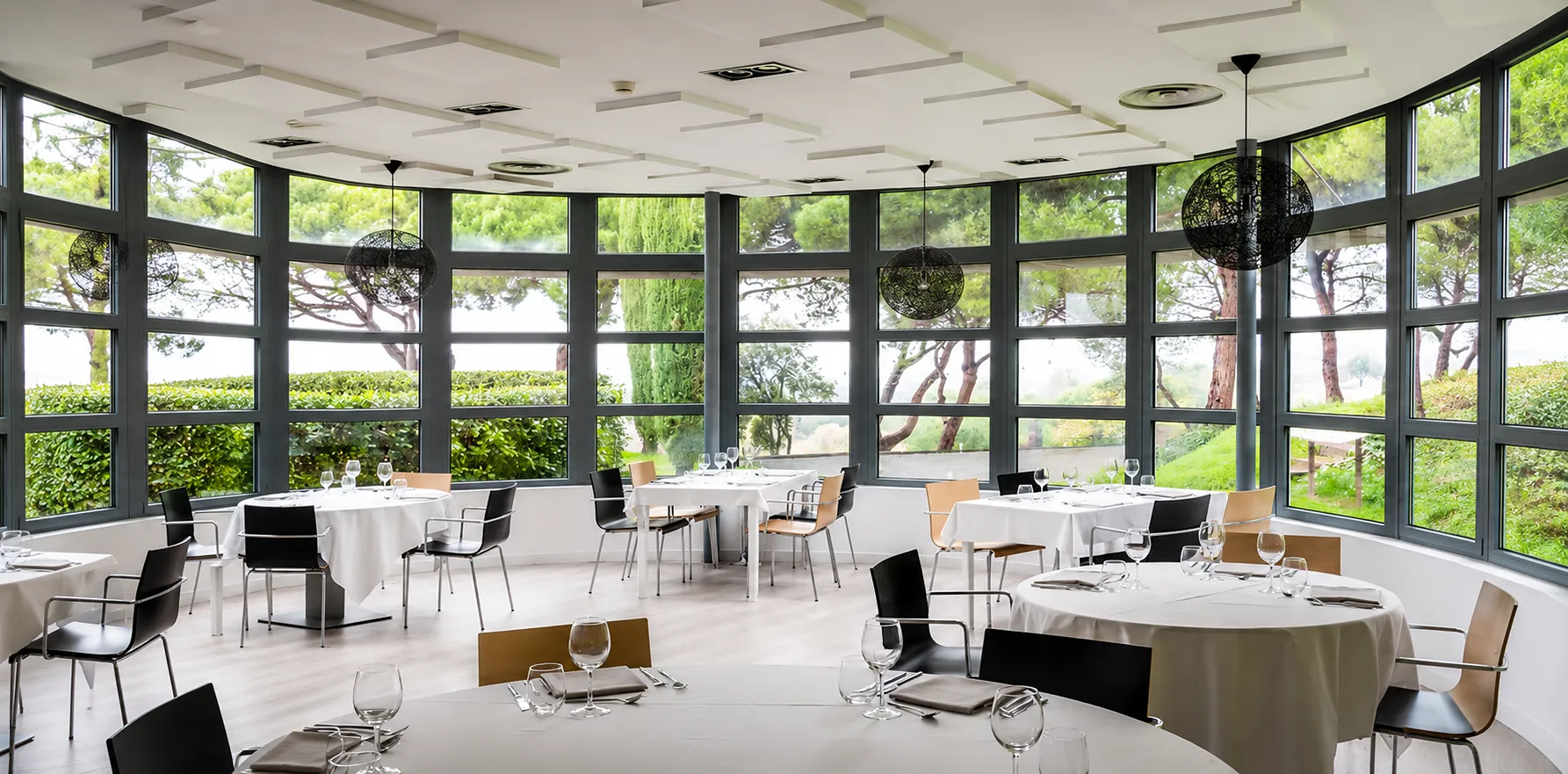 Comedor moderno con mesas servidas y ventanales con vista al jard&iacute;n, luz natural