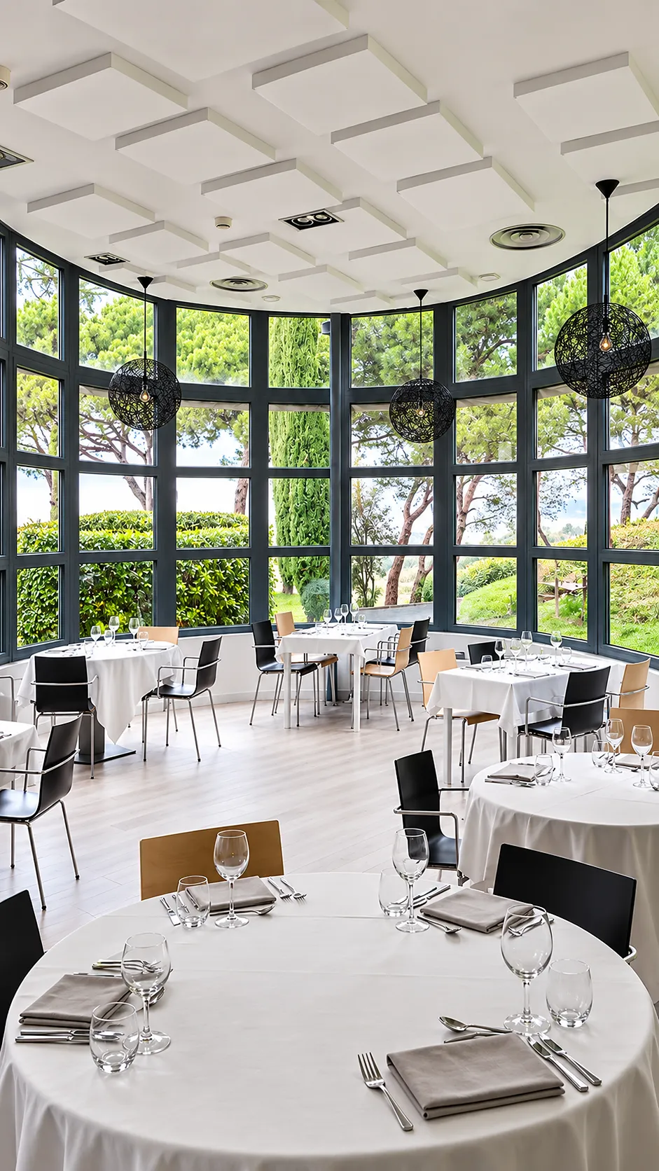 Comedor con mesas puestas, ventanales y l&aacute;mparas colgantes sobre jard&iacute;n verde.