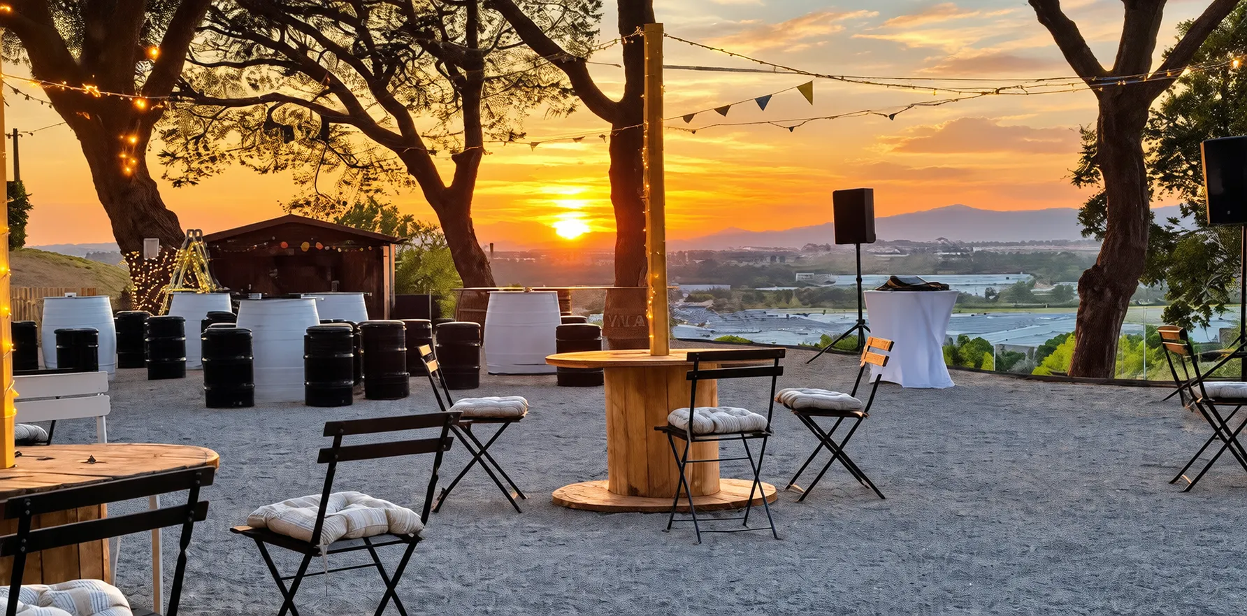 Terraza con sillas y mesa, luces y barriles; atardecer con lago al fondo.