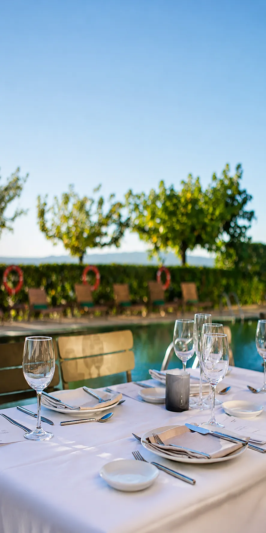 Mesa con copas y cubiertos junto a una piscina, cielo despejado y jard&iacute;n