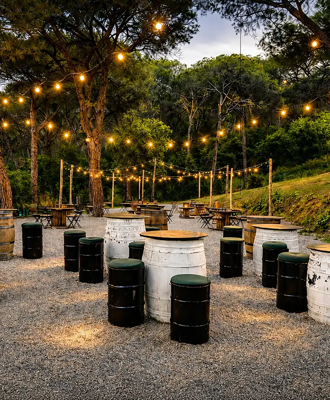 Espacio al aire libre con barriles y mesas, luces colgantes entre &aacute;rboles.