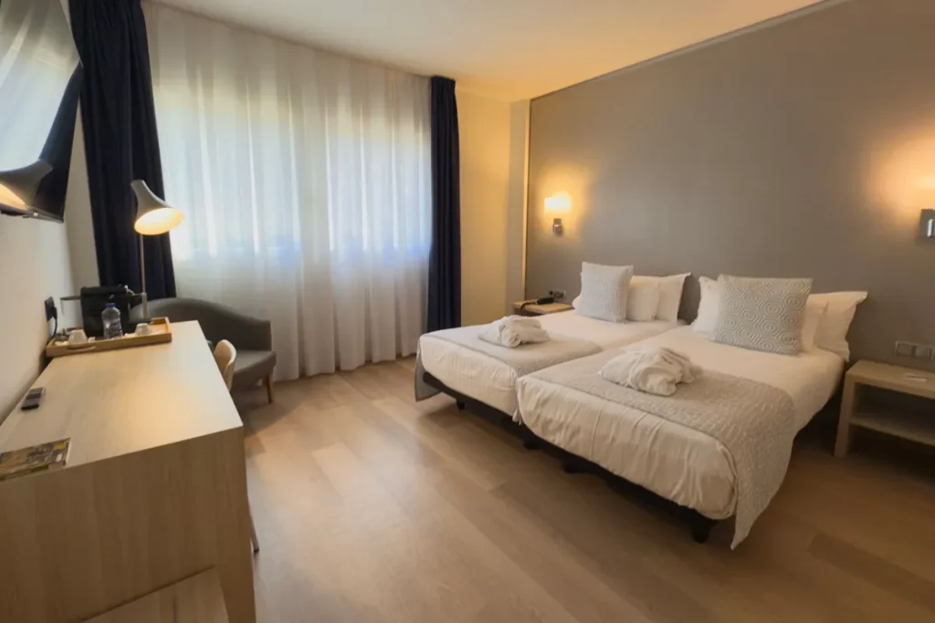 Habitaci&oacute;n de hotel con dos camas, cortinas y luces c&aacute;lidas al atardecer