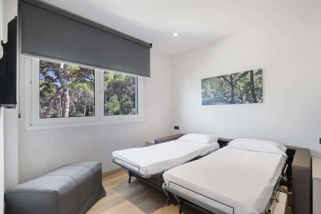 Habitaci&oacute;n blanca con dos camas, sof&aacute; gris y ventanas con vista al bosque