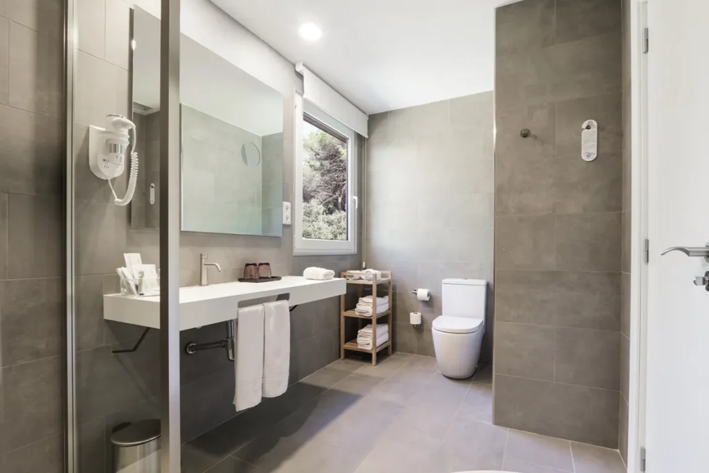 Ba&ntilde;o moderno con ducha de vidrio, lavabo, WC y ventana al exterior.