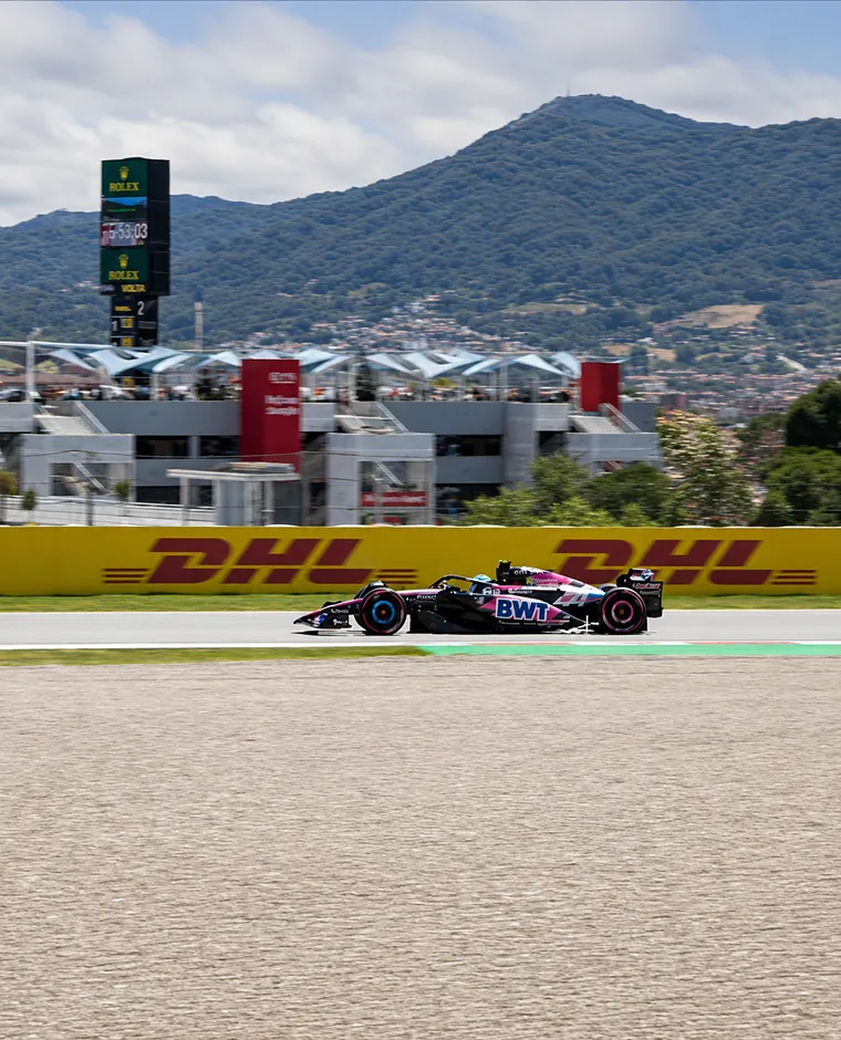 Auto de F&oacute;rmula 1 BWT en pista, con monta&ntilde;as y podio al fondo
