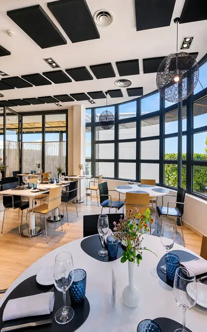 Sal&oacute;n moderno con mesas, copas y decoraci&oacute;n floral junto a ventanales.
