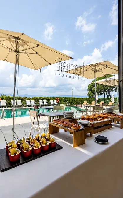 Mesa de buffet con c&oacute;cteles y aperitivos bajo sombrillas junto a piscina