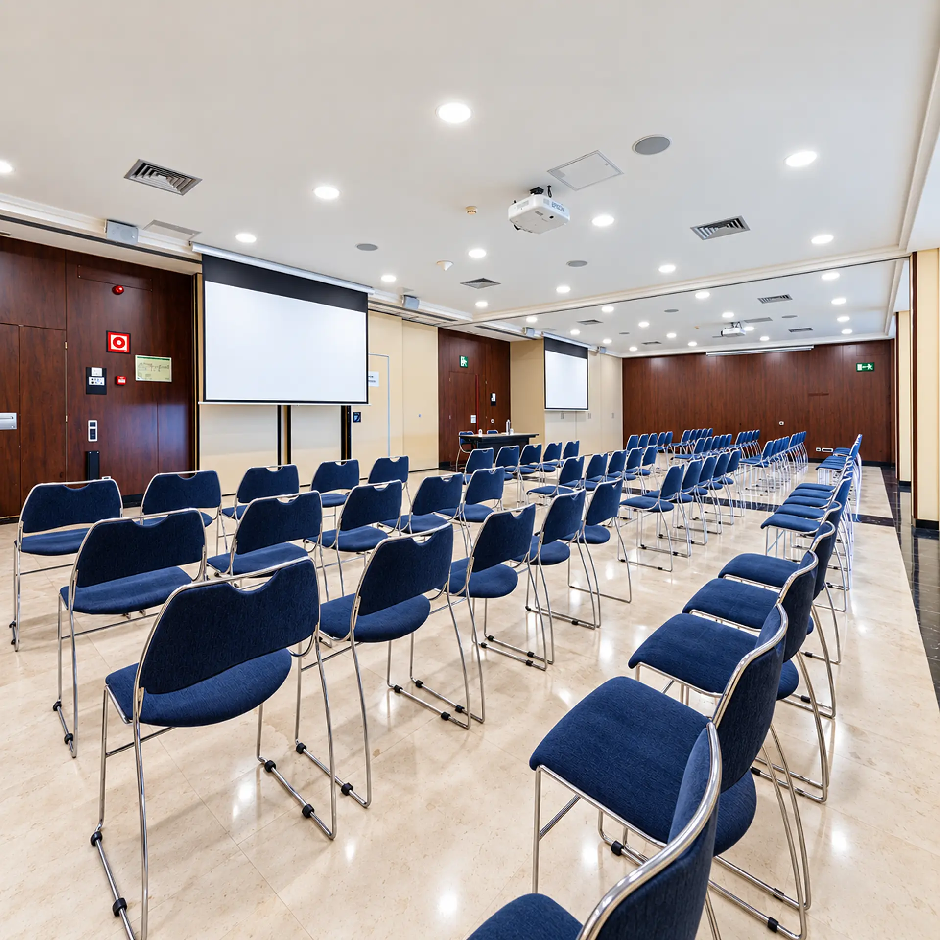 Sala de conferencias con sillas azules y pantallas de proyecci&oacute;n encendidas