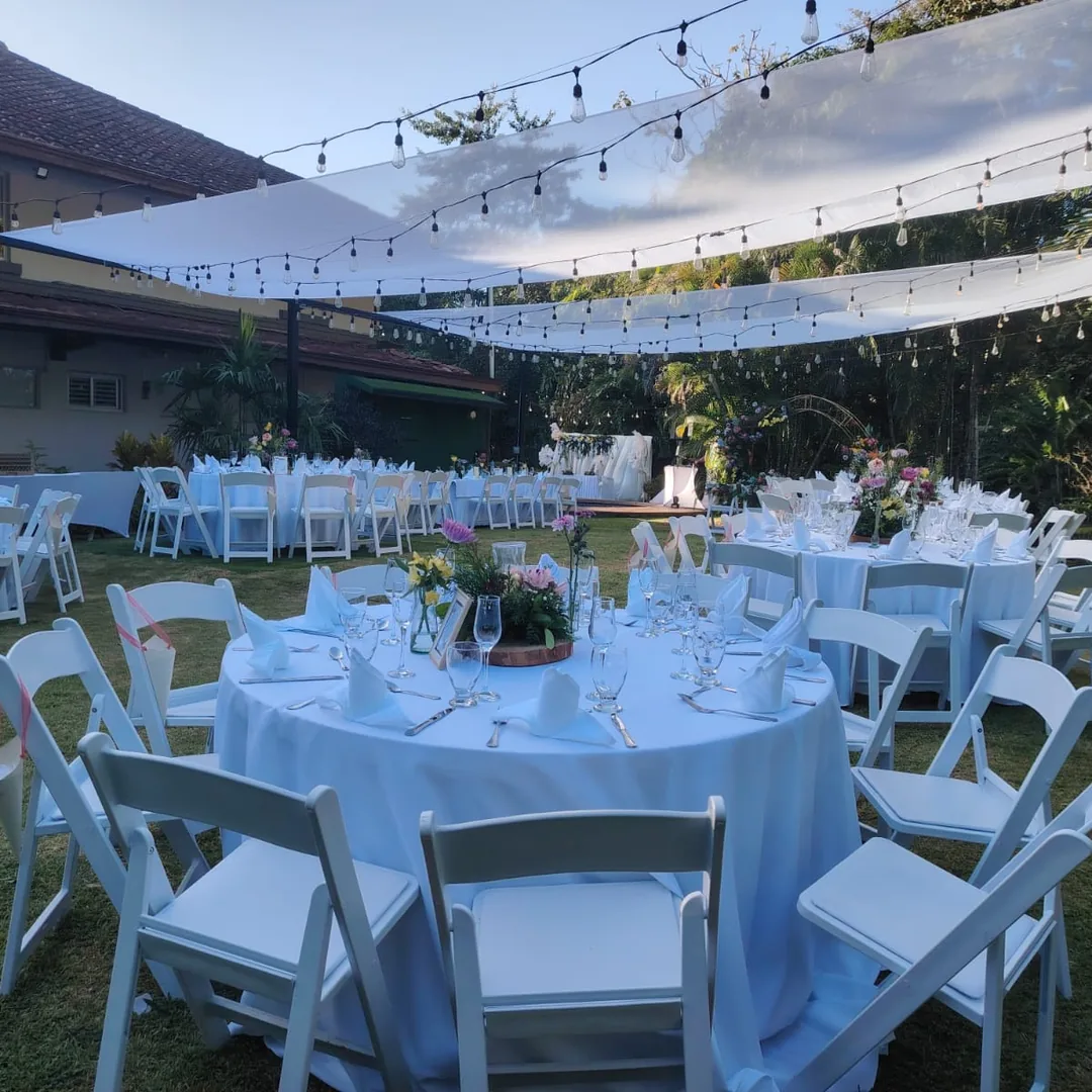 Mesa decorada con sillas blancas y guirnalda de luces en jard&iacute;n para evento al aire libre