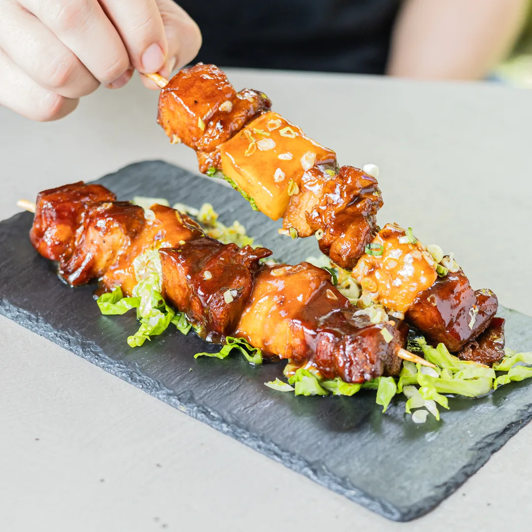 Mano sirviendo pinchos de pollo glaseados sobre piedra con lechuga y semillas