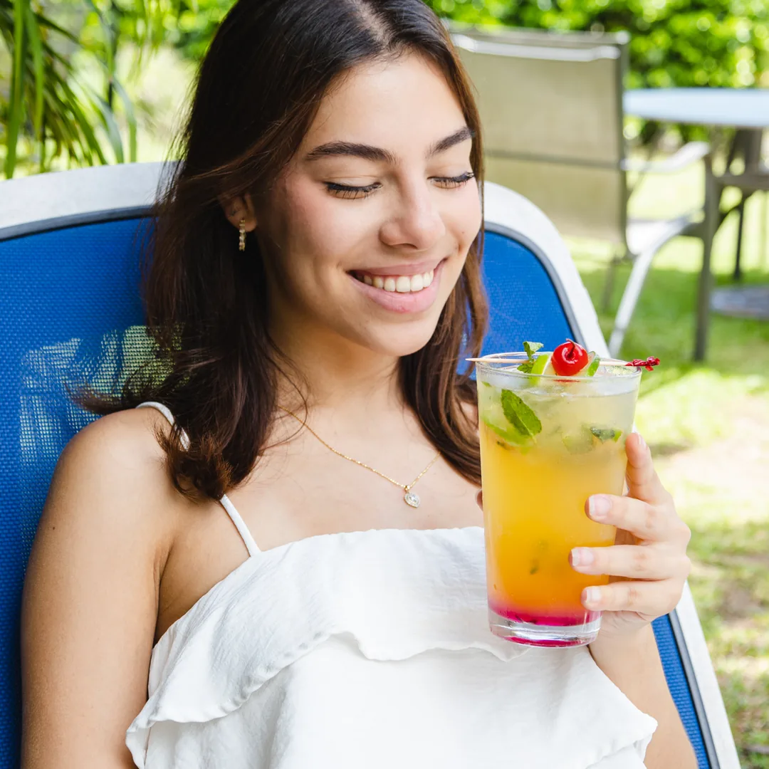 Mujer sonriente con c&oacute;ctel verde y rojo en jard&iacute;n, al aire libre.