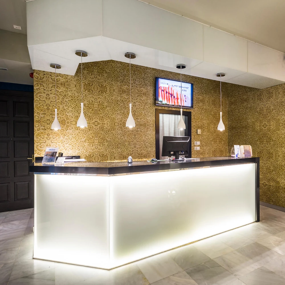 Recepci&oacute;n de hotel con mostrador blanco, l&aacute;mparas y pared con patr&oacute;n dorado