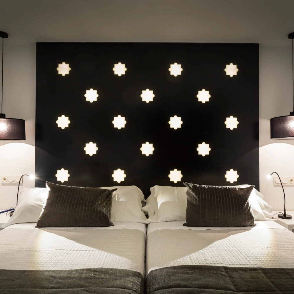 Cama doble con cojines blancos, luces de cruce decorativas en la pared oscura