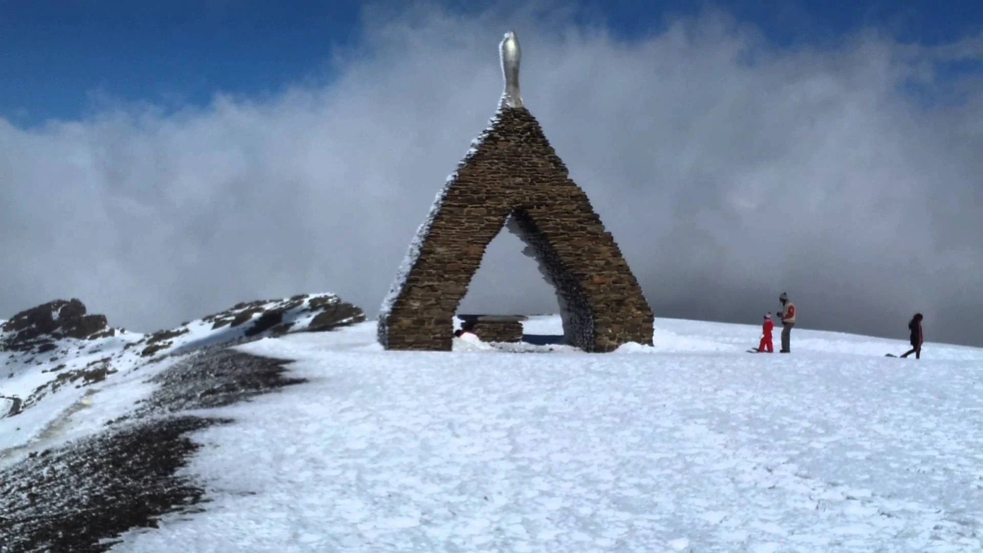 Escultura de piedra en forma de A, nieve y personas bajo el cielo nublado