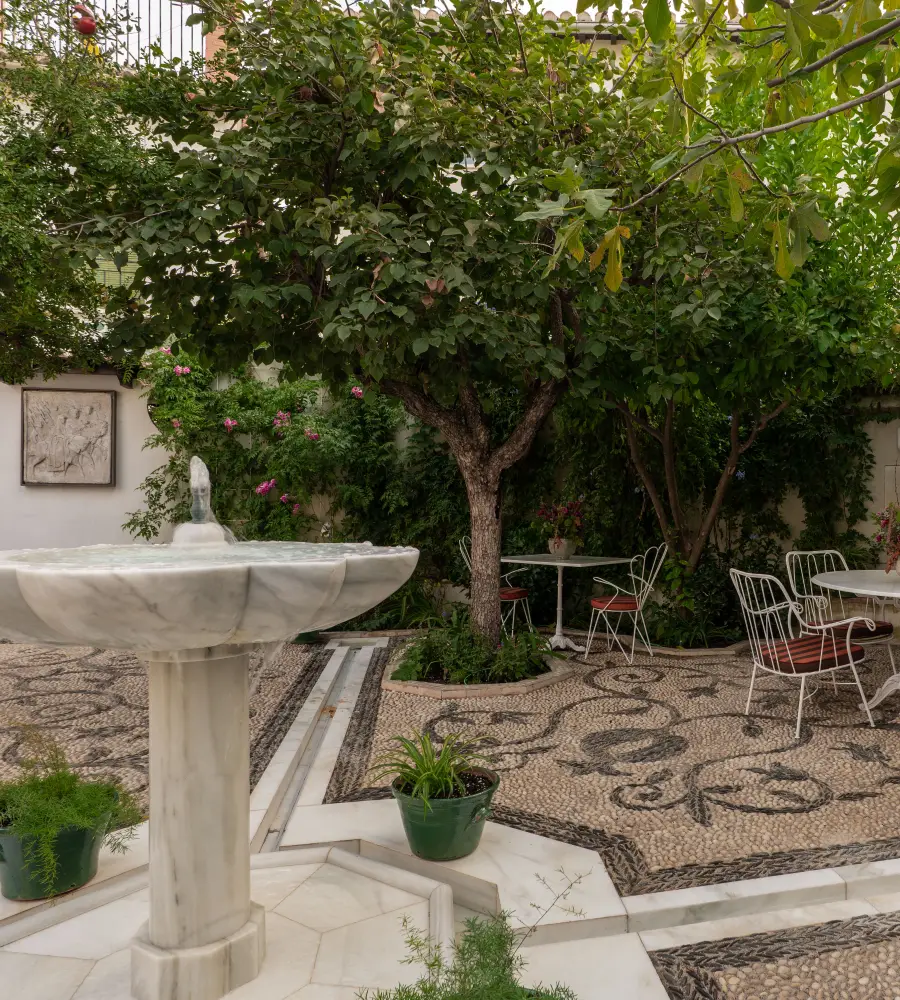 Patio con fuente de piedra, jard&iacute;n verde y sillas met&aacute;licas en el suelo empedrado
