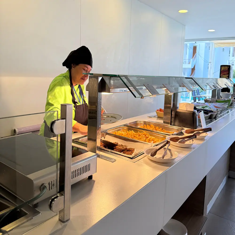 Cocinera sirve comida en l&iacute;nea de buffet con recipientes y verduras.