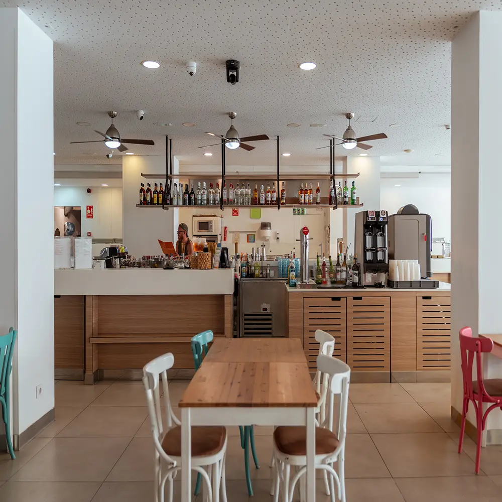 Bar con barra de bebidas, cafeteras y mesas con sillas en un local interior