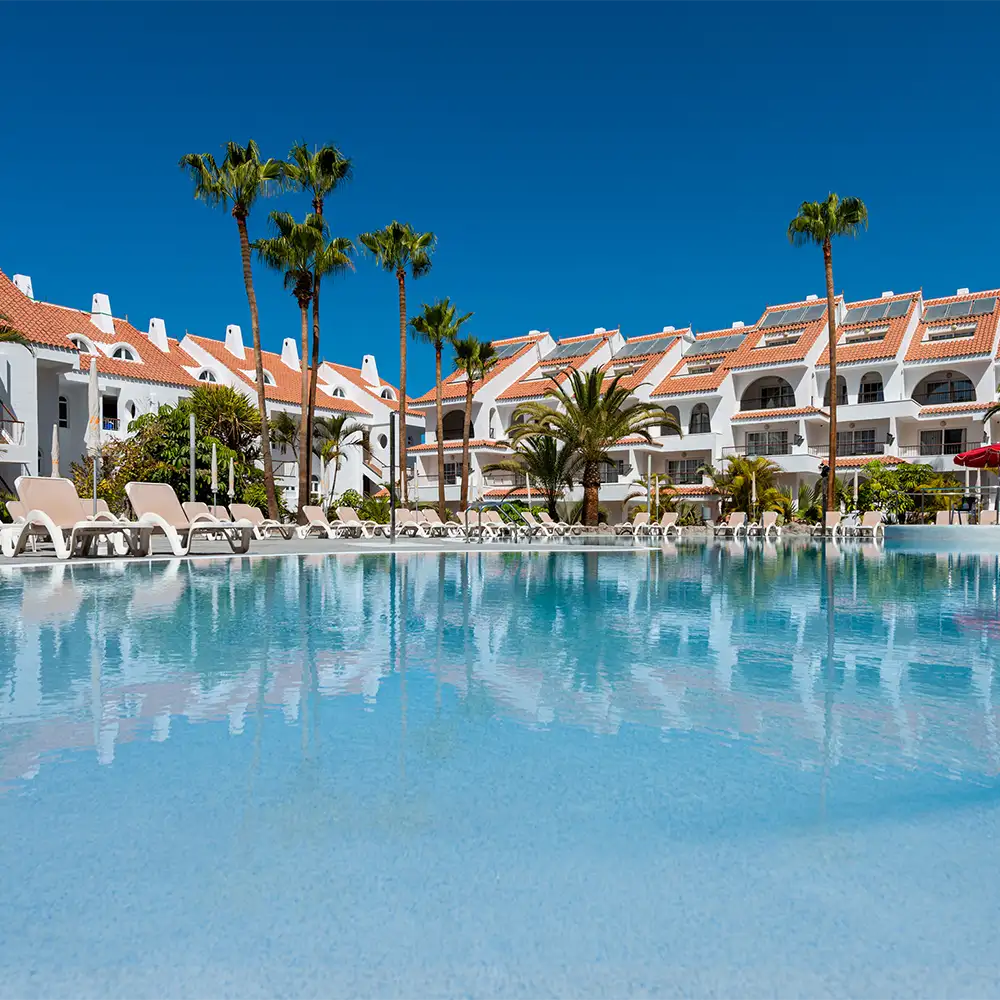 Piscina y palmeras frente a caba&ntilde;as blancas con techo de tejas rojas.