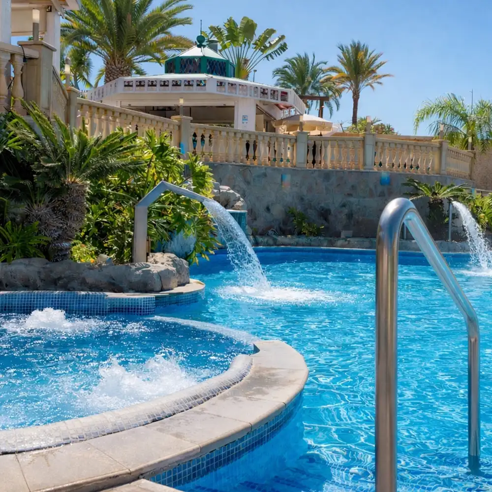 Piscina tropical con tobog&aacute;n y palmeras, bajo cielo azul.