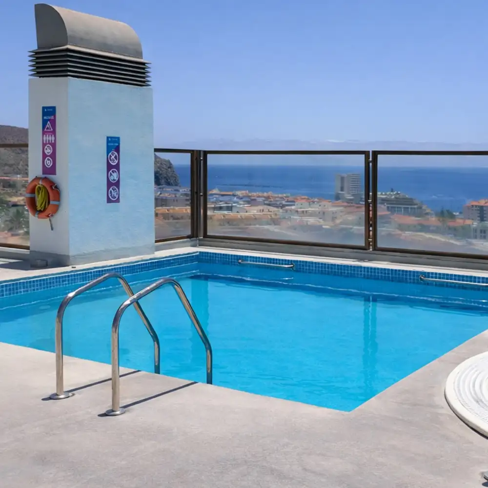 Piscina azul con vistas al mar desde un hotel, junto a barandillas met&aacute;licas.