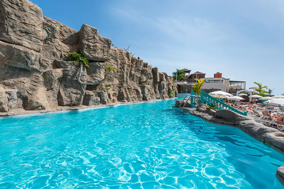 Piscina azul junto a rocas y tumbonas, con edificios y cielo despejado al fondo