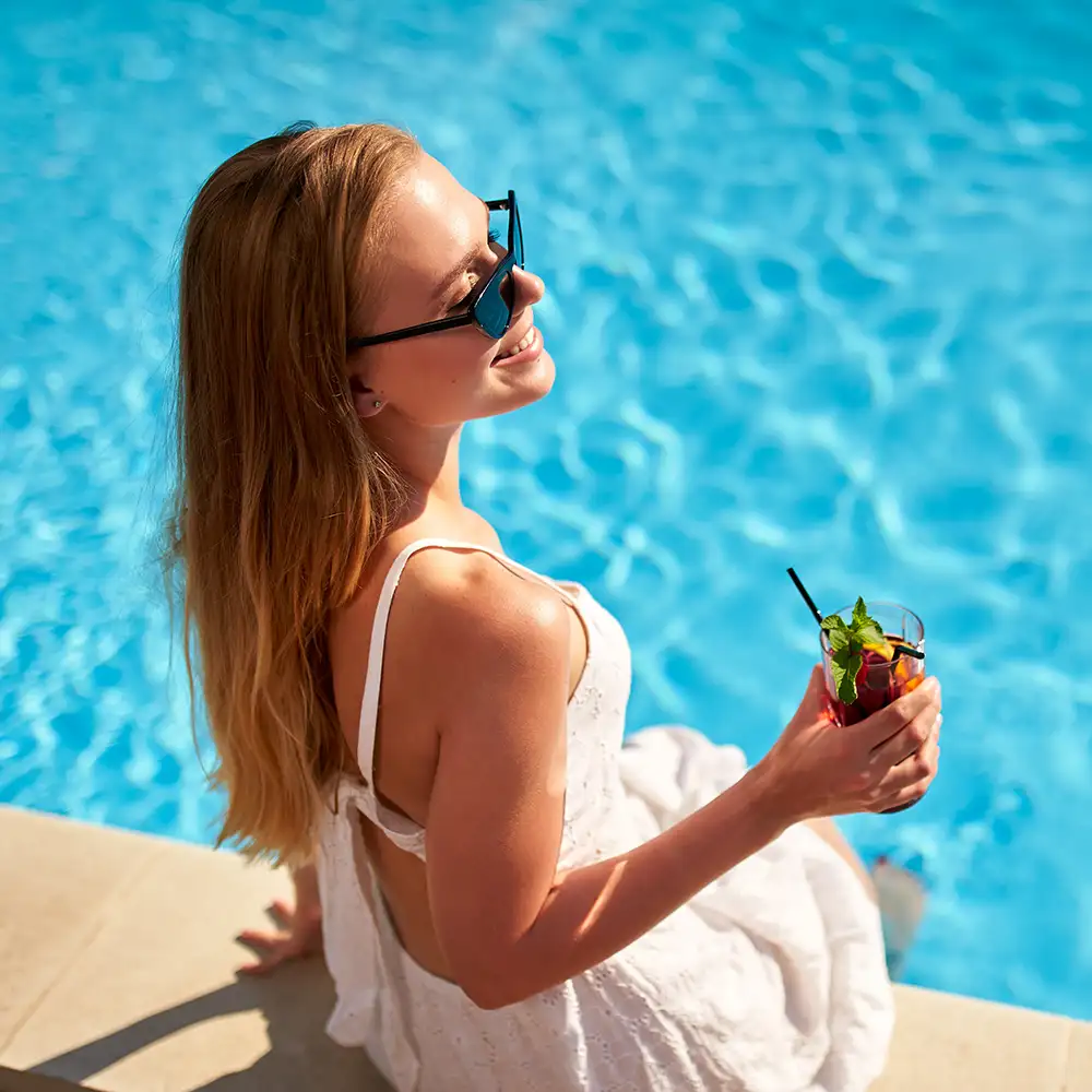 Mujer con gafas de sol tomando un c&oacute;ctel junto a una piscina.