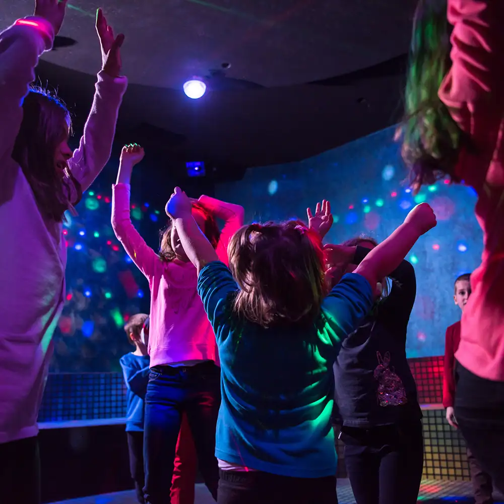 Ni&ntilde;os bailando en una fiesta con luces de colores y DJ en el fondo.