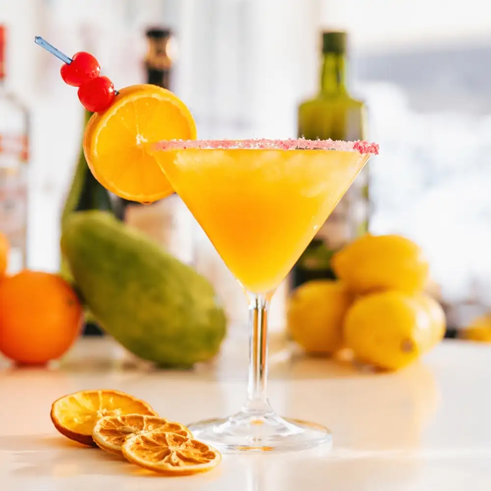 C&oacute;ctel de naranja con borde rosado, aceitunas y frutas sobre mesa blanca