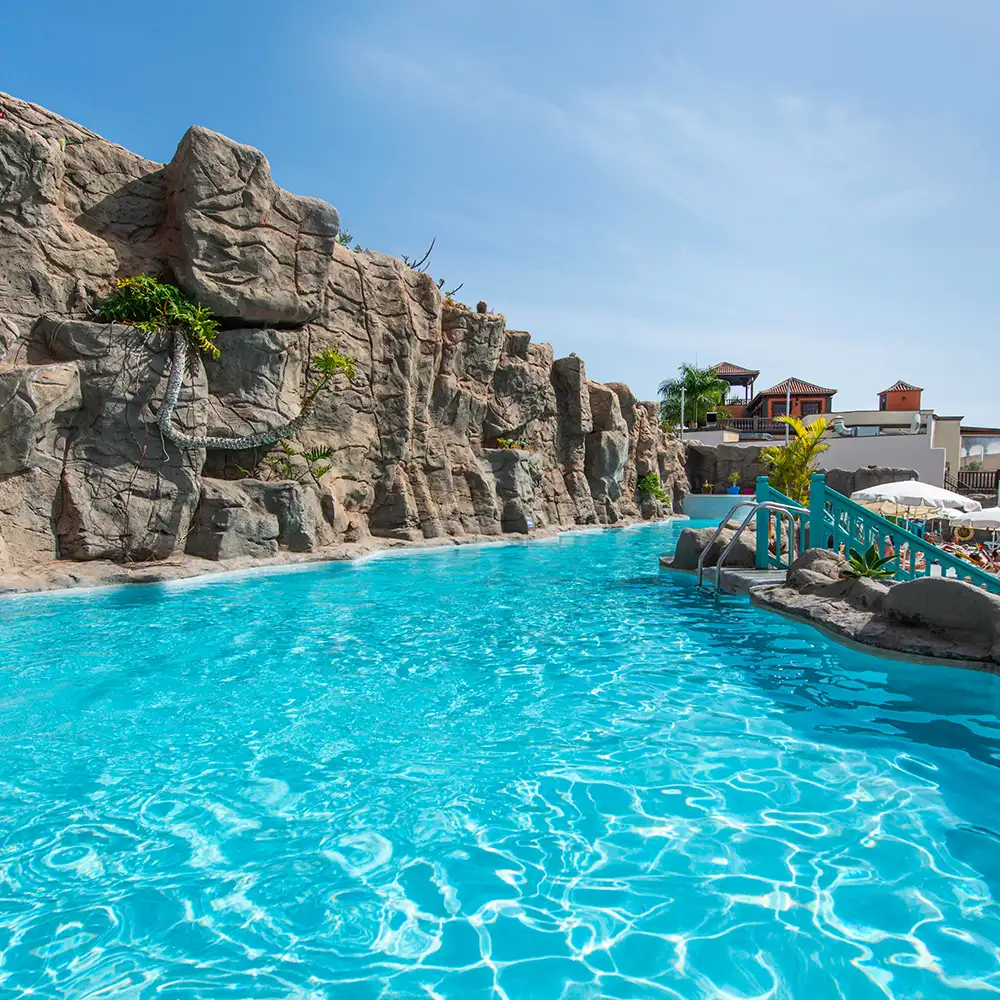 Piscina turquesa con rocas y barandilla azul, junto a casas al fondo