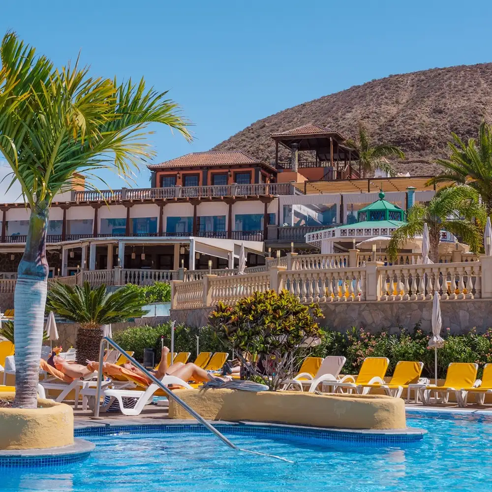 Piscina con tumbonas y palmeras frente a hotel junto a colina y cielo azul