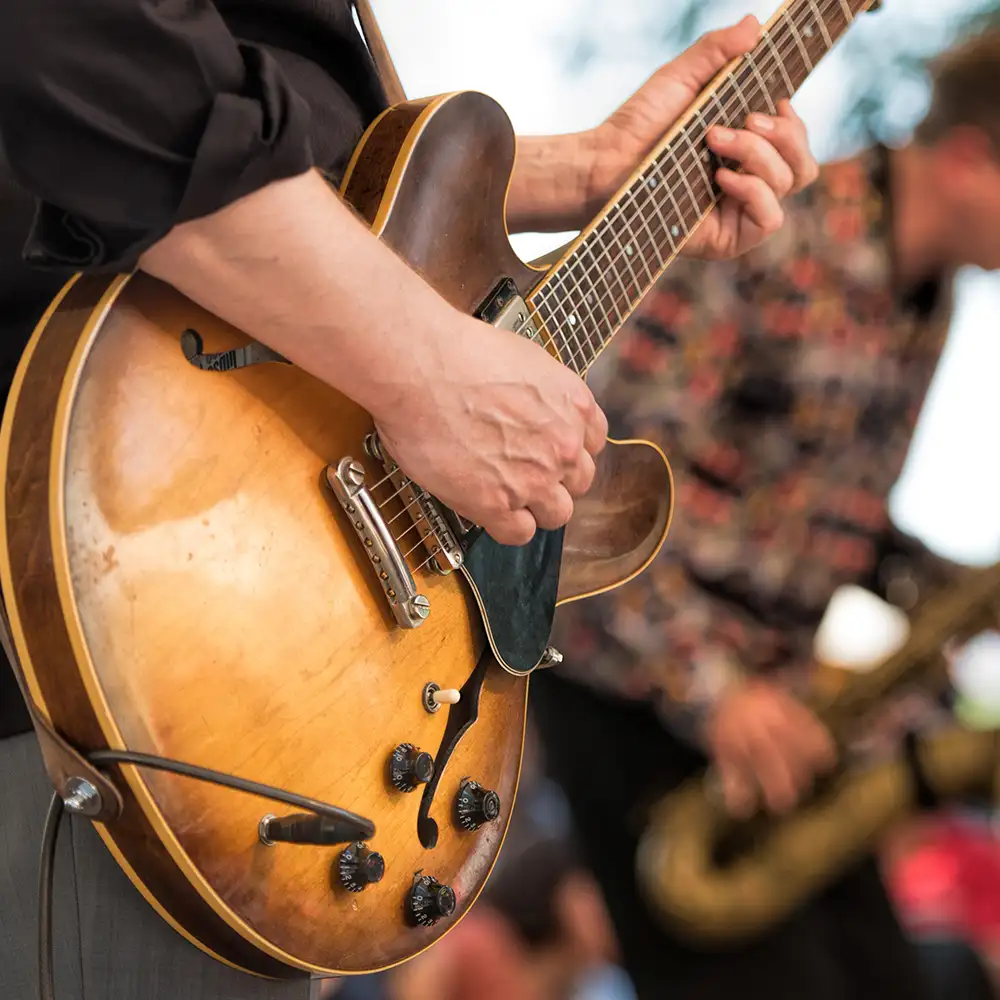 Mano afinando una guitarra ac&uacute;stica en un escenario con p&uacute;blico desenfocado