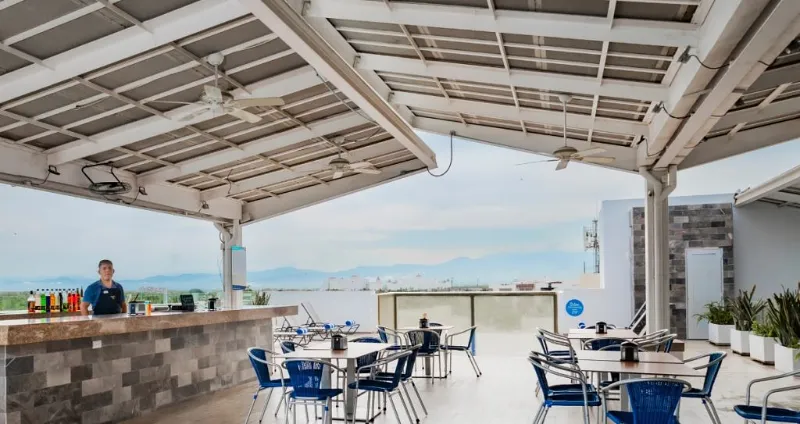Terraza con mesas y sillas bajo techo, vista al paisaje y a la ciudad.