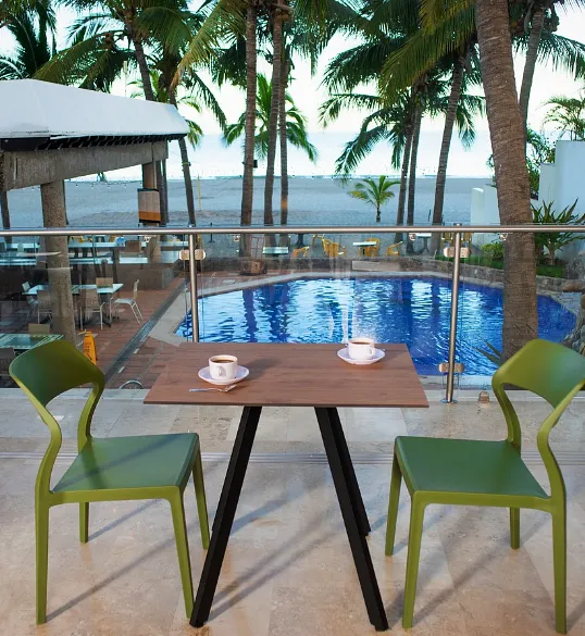Mesa con dos tazas y sillas verdes frente a piscina y palmeras al mar
