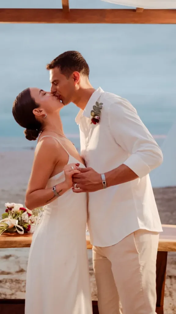 Pareja en boda bes&aacute;ndose frente al mar, el novio con camisa blanca y ella vestido blanco