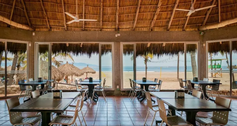 Comedor al aire libre con mesas y vista al mar bajo techo de palma